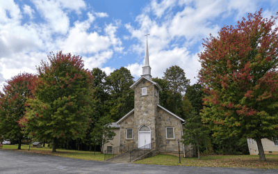 Small stone church with a white steeple and steps, set among autumn-colored trees under a blue sky.