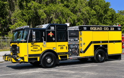 Yellow fire rescue engine labeled squad co. 23, with open tool compartments, parked in a lot with trees in the background.