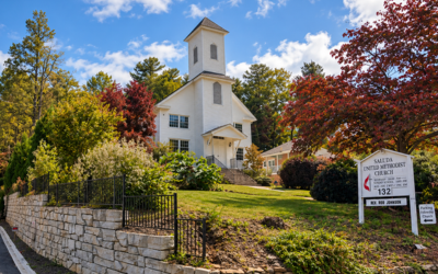 White church with a tall steeple, set among colorful autumn trees on a sunny day.