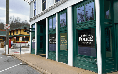 Exterior of a green-framed storefront police building along a sidewalk, with 'saluda police' signage in the windows and a no-left-turn sign nearby.