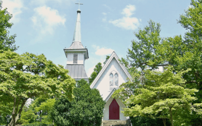 Small white church with a tall steeple and red front door, surrounded by green trees on a sunny day.
