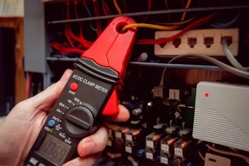 “Hand holding an AC/DC clamp meter around a yellow wire inside an electrical control panel with multiple cables and components.”