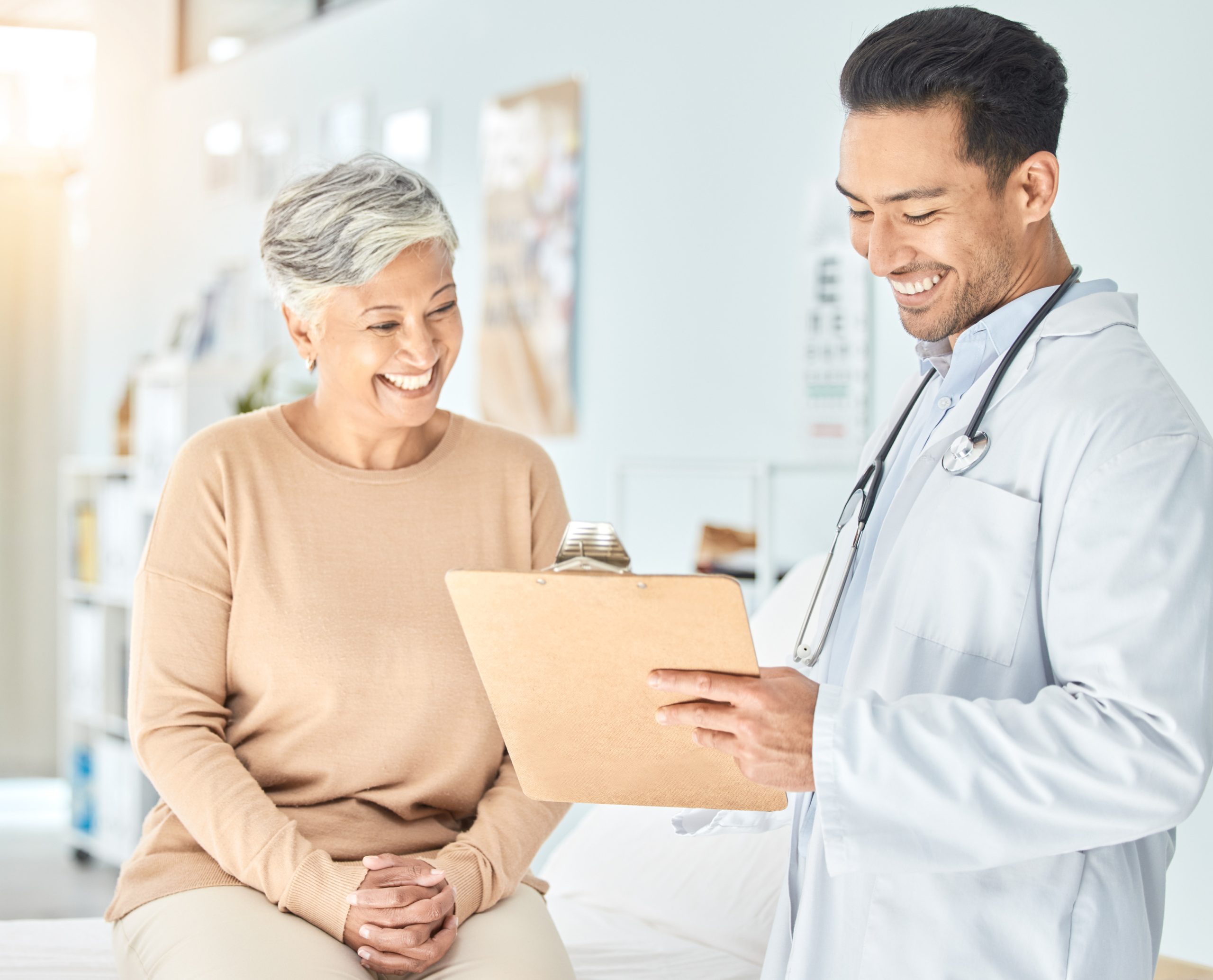 A smiling doctor talks goes over a chart on a clipboard with a smiling female patient in a brightly lit room.