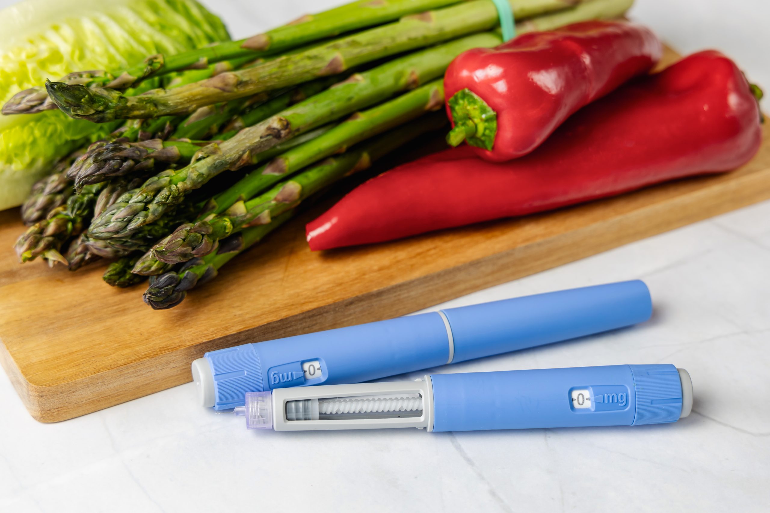 A GLP-1 injector, like Wegovy, Zepbound, or Ozempic, alongside a cutting board with health foods like peppers, asparagus and lettuce.