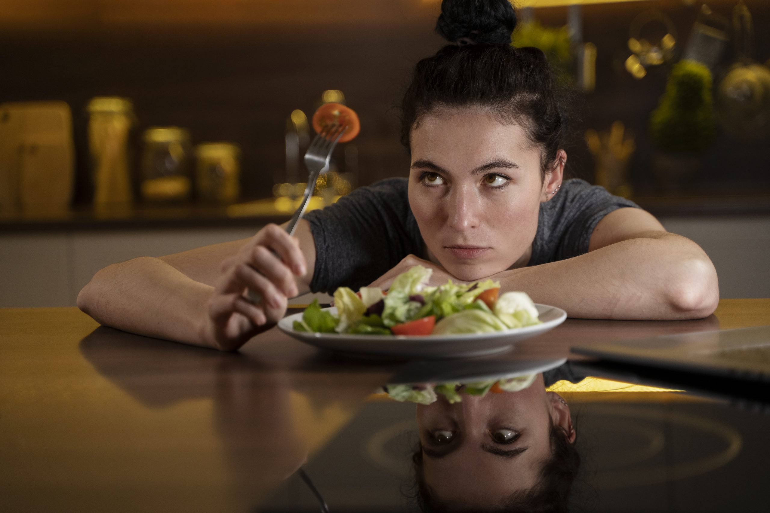 A woman looks incredulously at a tomato on a fork. She has her chin rested on her arm on a table with a salad in front of her.