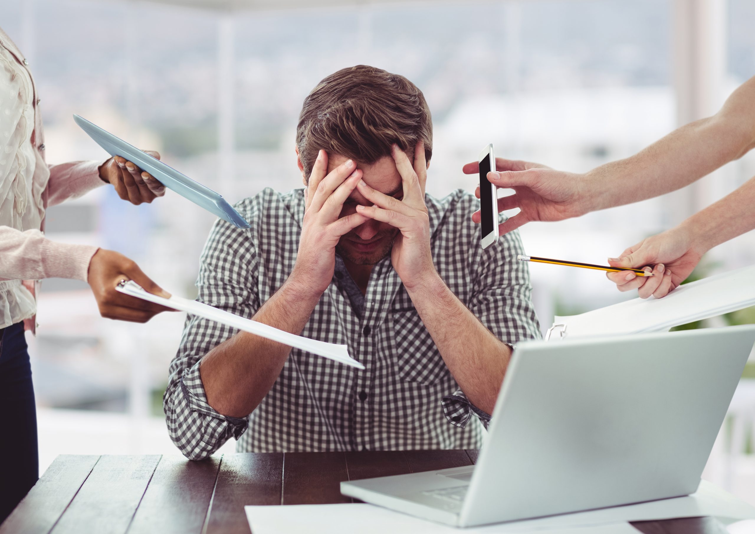 A man has his hands over his face. He looks stressed. He has a computer on a desk in front of him. People off-screen are handing him things like papers, tablets, phones, and pencils.
