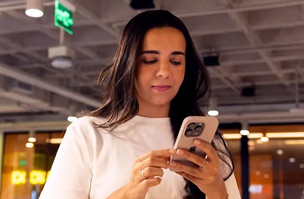 ulher jovem com cabelos escuros ondulados e camiseta branca olha focada para a tela do seu smartphone em um escritório moderno com iluminação industrial suave ao fundo.ulher jovem com cabelos escuros ondulados e camiseta branca olha focada para a tela do seu smartphone em um escritório moderno com iluminação industrial suave ao fundo.