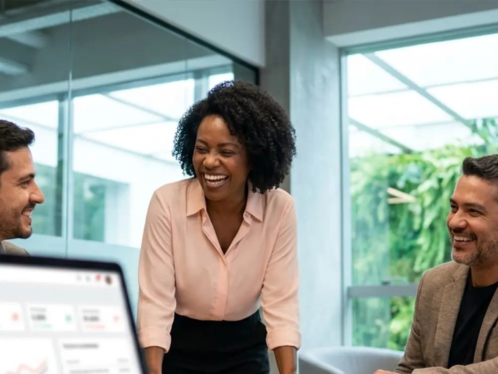 Mulher negra com blusa rosa claro sorri alegremente ao lado de dois colegas em sala de reuniões com paredes de vidro. Um laptop exibindo um dashboard de métricas está em primeiro plano desfocado.