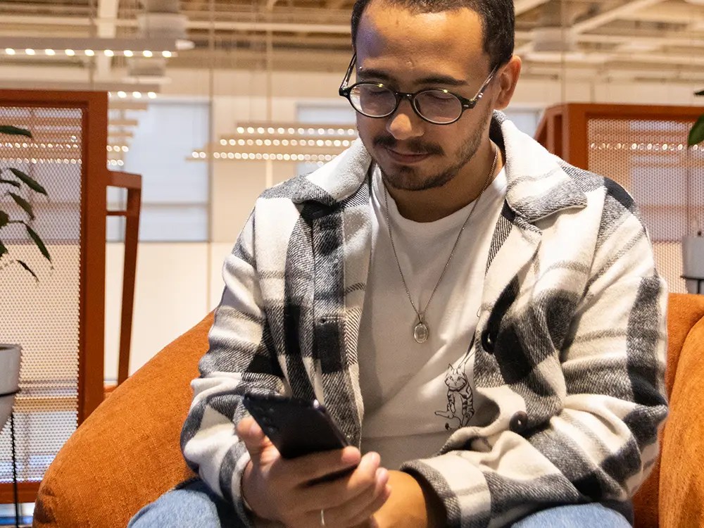 ovem de óculos e casaco xadrez cinza e branco está sentado em uma poltrona laranja confortável, sorrindo sutilmente enquanto digita no celular.