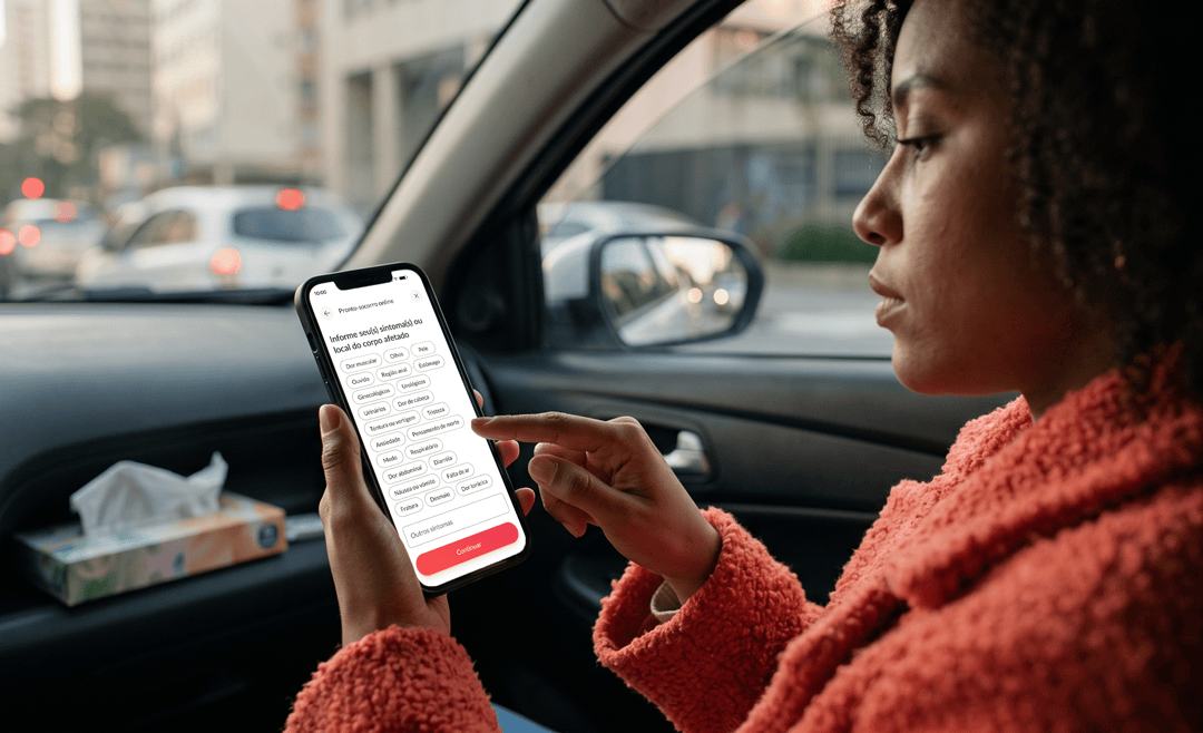 Mulher de casaco coral no banco de passageiro de carro selecionando seus sintomas de dor ou mal-estar na tela de pronto-socorro online do aplicativo da Sami.