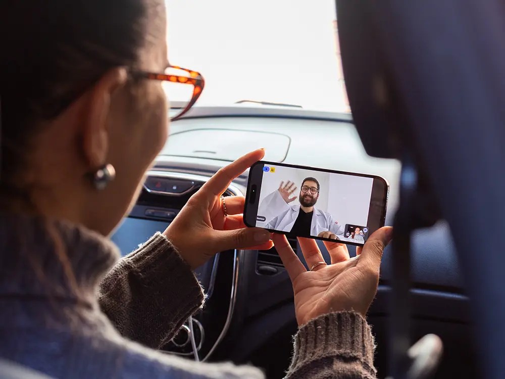 isão de trás de mulher com óculos no interior de um carro realizando teleconsulta no celular com médico de família barbudo que acena sorridente vestindo jaleco branco.