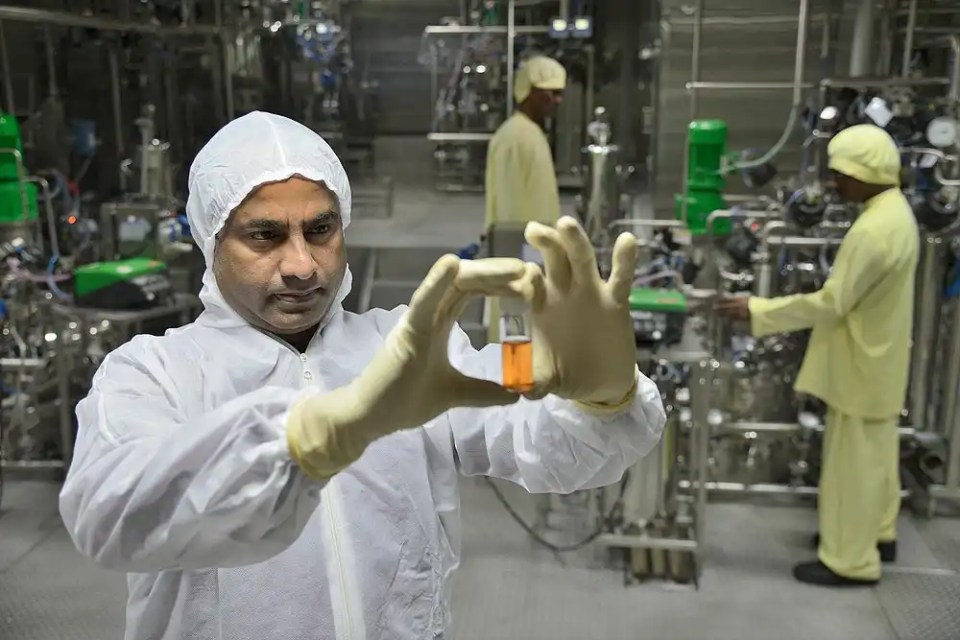 A scientist in a protective suit examines a vaccine vial in a laboratory setting, with other personnel working in the background.