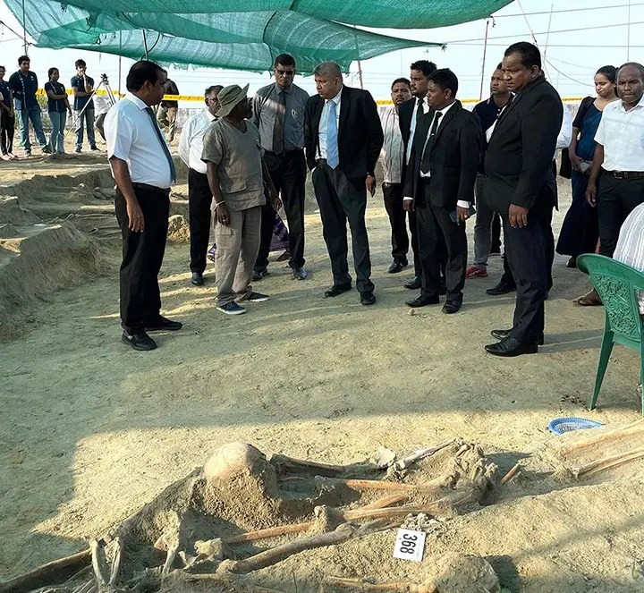 Group of officials in suits and workers gathered around an open excavation pit, examining partially exposed human bones under a green shade at an archaeological/dig site.