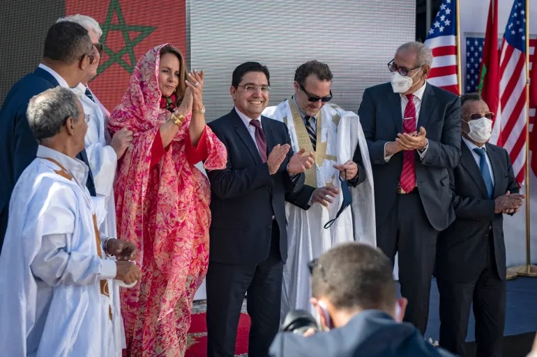 Group of dignitaries on a stage applauding, with a woman in pink traditional dress and men in suits; Moroccan and U.S. flags in the background.