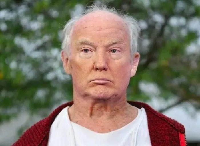 Older man with white hair and pale skin, wearing a white shirt and red cardigan, outdoors with greenery behind him, neutral expression