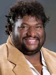 Portrait of a smiling man with dark, curly hair and a beard, wearing a tan jacket over a white shirt.