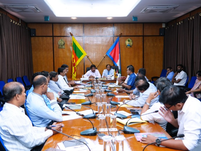 Group of officials around a long conference table in a formal meeting room with microphones and documents.