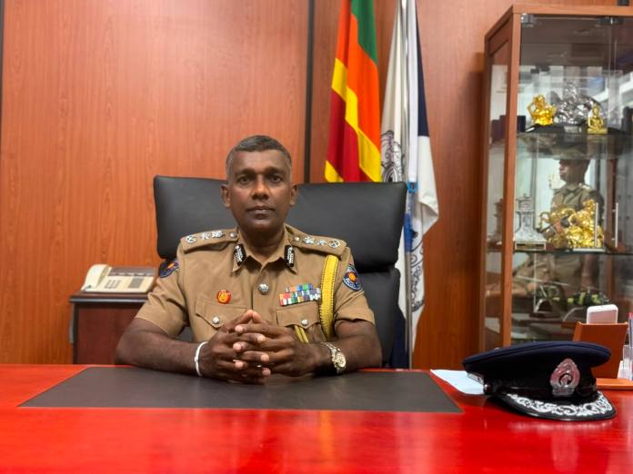 Policer officer in a khaki uniform seated behind a red desk in an office with flags and a glass display cabinet in the background.