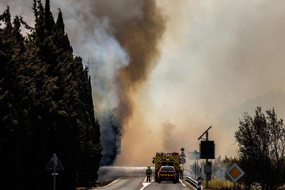 Emergency response on a rural road as thick smoke rises from a fire; firefighters and a striped fire truck block the lane.