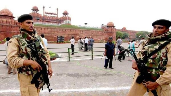 Two armed soldiers in camouflage guard outside the Red Fort, with visitors in the background.