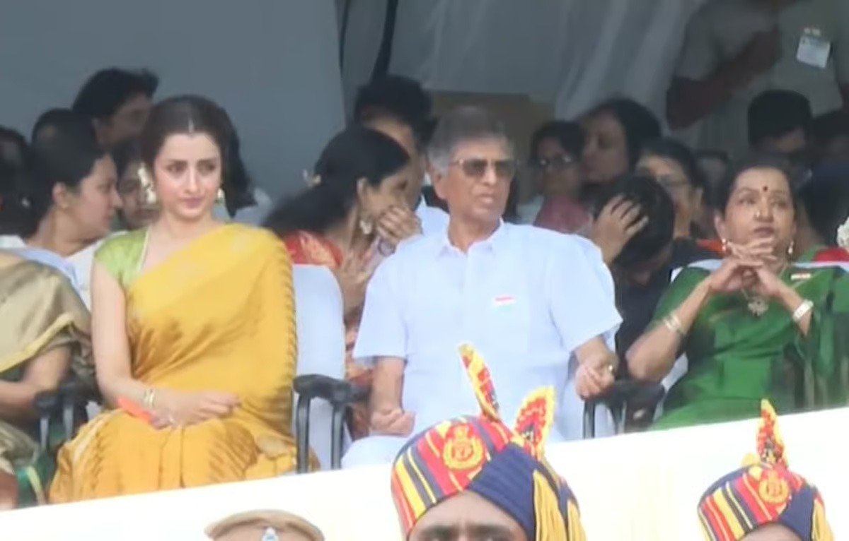 Three people seated in a crowd at a daytime event: a woman in a yellow sari, a man in a white kurta with sunglasses, and a woman in a green sari on the right.