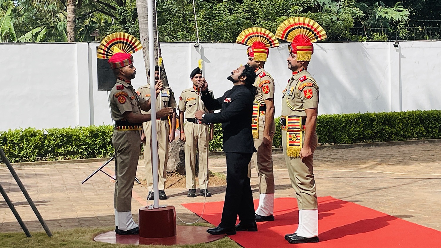 A man in a black suit hands a flag to a uniformed soldier during a flag-raising ceremony, with other guards standing at attention on a red carpeted area.