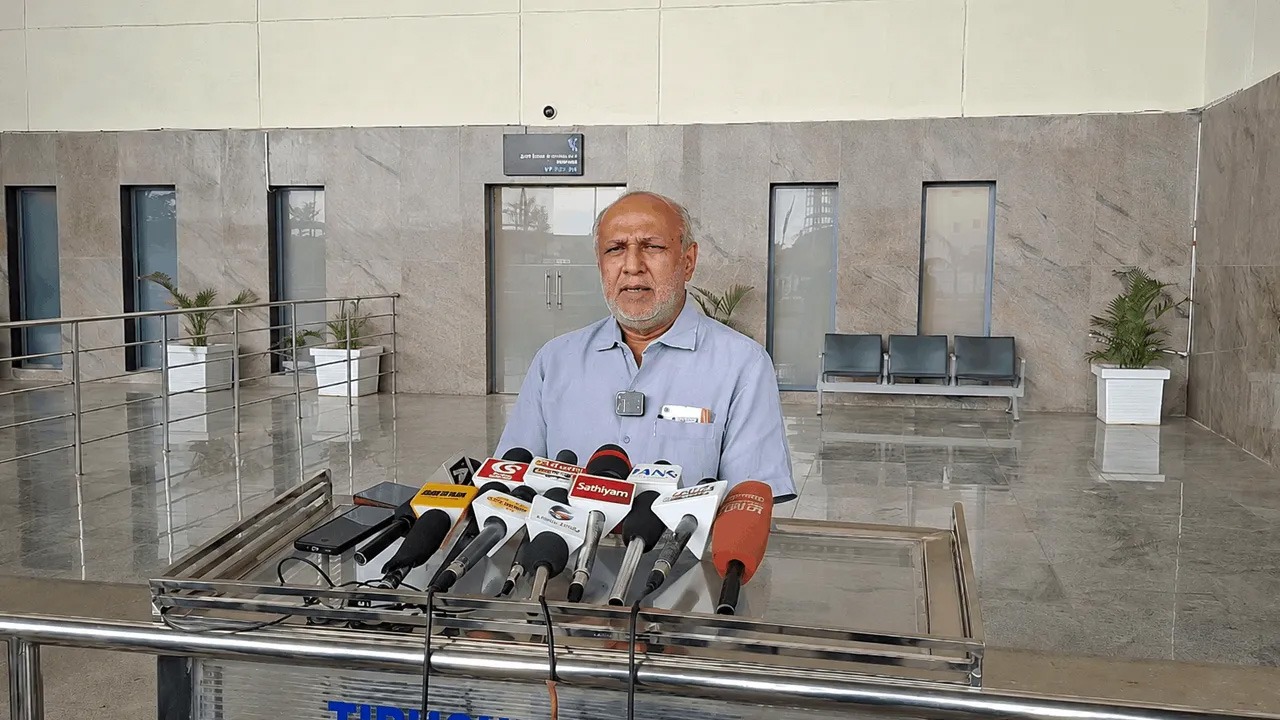 Older man in a light blue shirt stands at a metal podium with many microphones, addressing reporters in a marble-walled lobby.