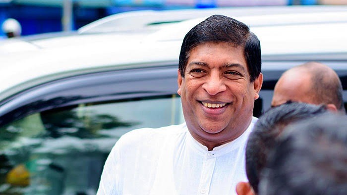 Smiling man in a white kurta standing beside a car, surrounded by people outdoors.