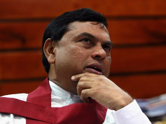 Close-up of a man wearing a white shirt and red ceremonial sash, resting his chin on his hand in a contemplative pose against a warm background.