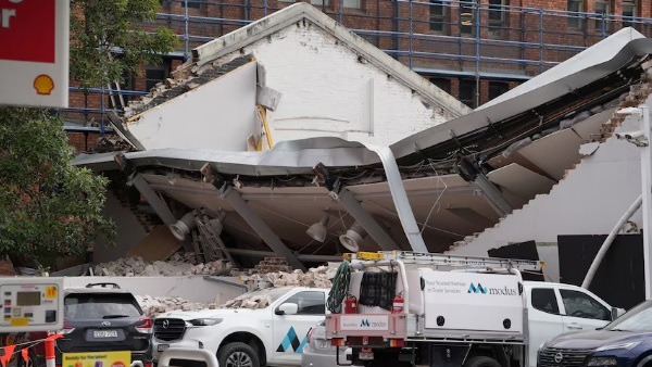 Building partially collapsed with exposed rubble and toppled roof, with emergency response vehicles and a work van parked in front on a city street.