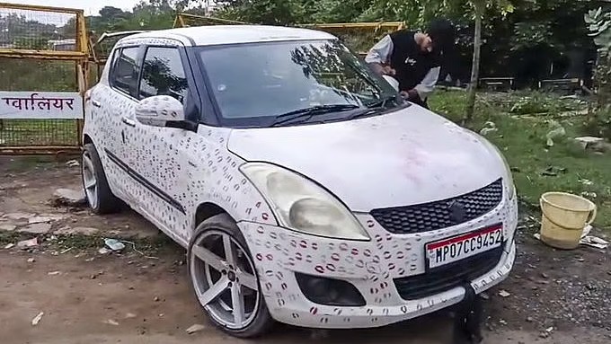 White hatchback parked on a dirt lot with a dotted camouflage wrap; Indian license plate MP07CC9452, and a man stands near with a yellow bucket nearby.