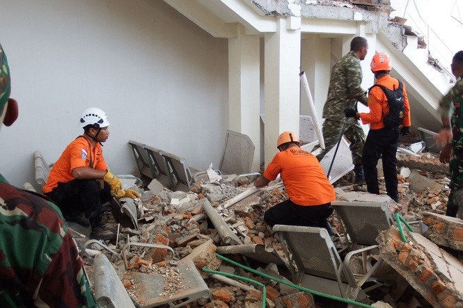 Rescue workers in orange uniforms and helmets sift through rubble and broken chairs beside a damaged building.