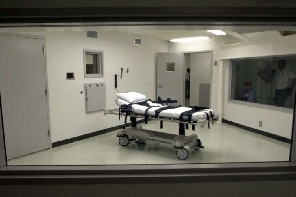 Hospital room with a wheeled stretcher secured with straps and white sheets, viewed through a glass window from outside the room.