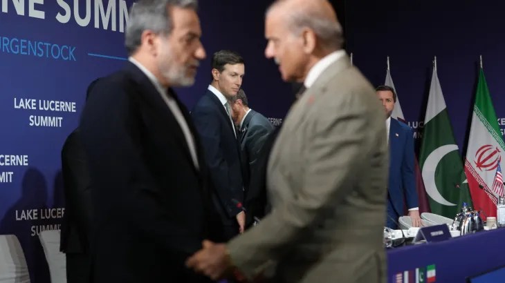 Businessmen shake hands in the foreground at a Lake Lucerne Summit event, with a panel and flags in the background.