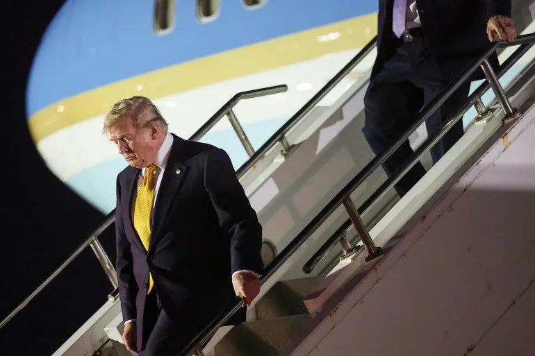 Man in a dark suit and yellow tie descending airplane stairs at night with a large jet in the background.