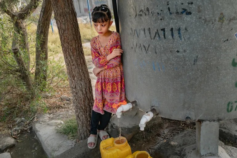 Girl in a colorful dress stands beside an outdoor water tank, watching water drip into a yellow container below the taps.