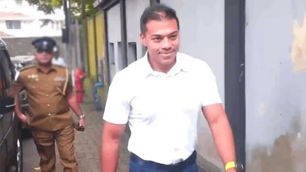 Man in a white polo shirt walks toward the camera along a narrow street, with a uniformed officer in brown in the background.