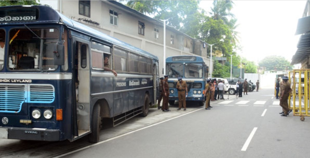 Police officers standing near blue buses parked along a street, with a crosswalk and white building in the background.