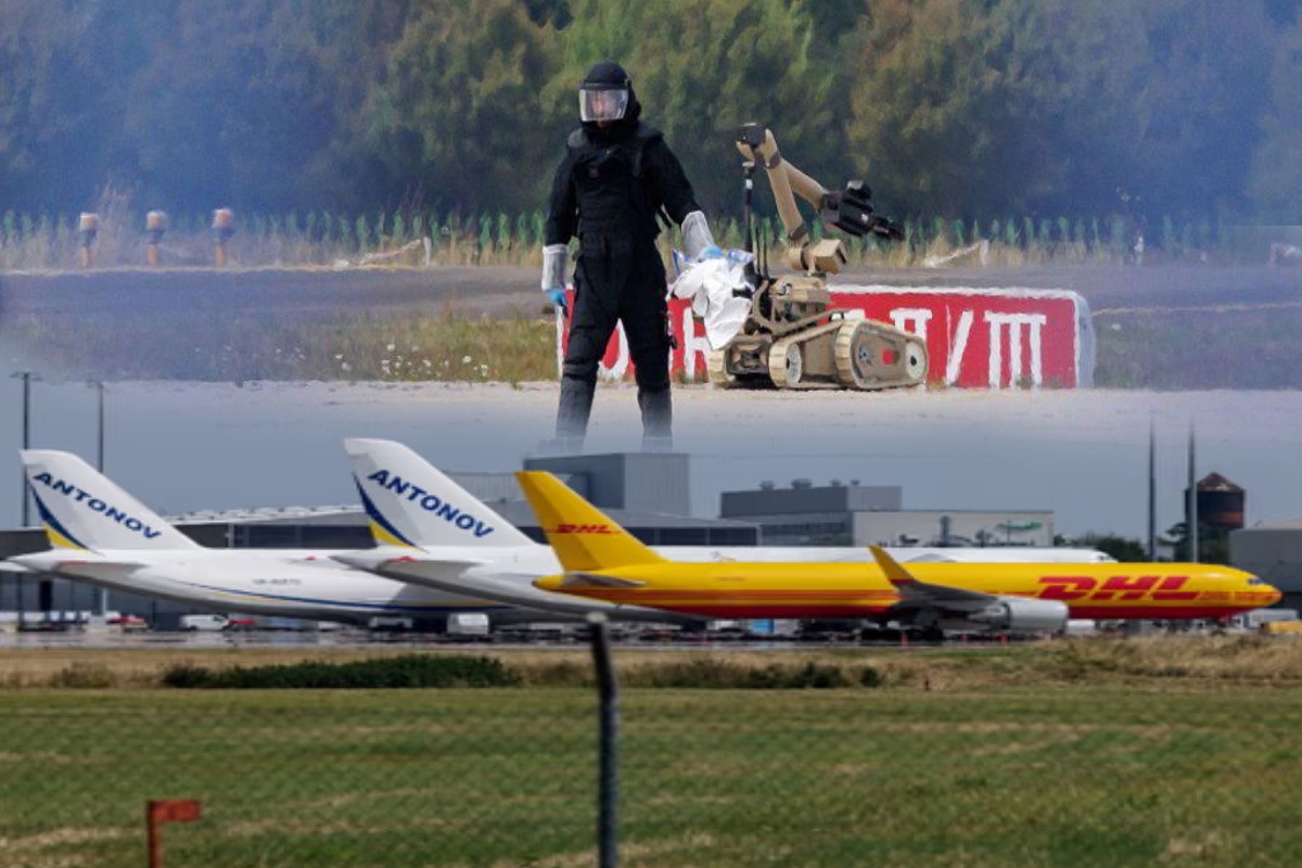 Person in a black hazmat suit and helmet stands beside a ground robot on an airfield with cargo planes in the background (Antonov, DHL).