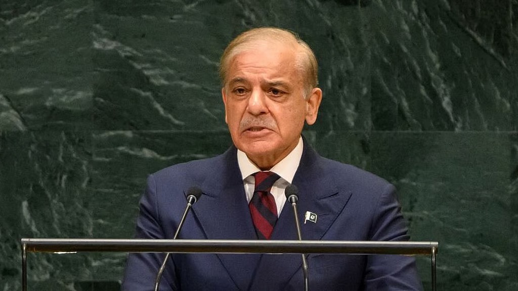 Older man in a navy suit speaks at a podium with two microphones against a green marble backdrop.