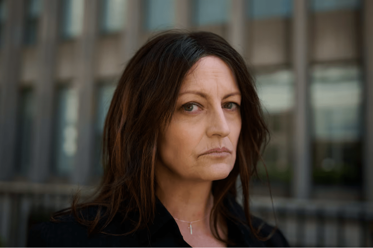Serious-looking woman with brown hair in a black top outdoors in front of a glass building.