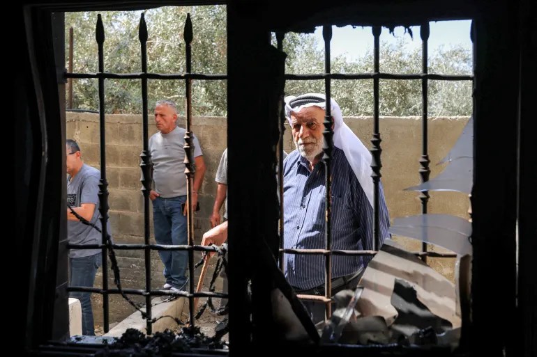Three men stand outside a burned building, seen through a shattered window with iron bars and debris inside.