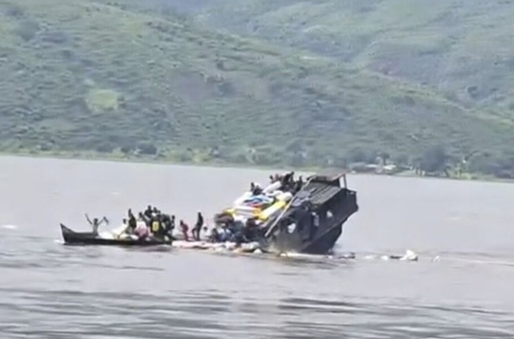 A capsized vessel on a calm lake, with people in a small rescue boat approaching and others clinging to the hull, green hills in the distance.
