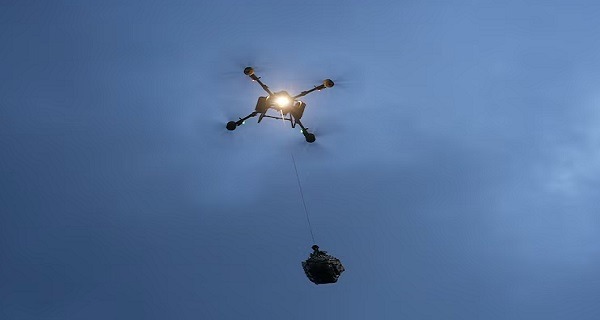Quadcopter drone with four arms lifting a payload under a clear blue sky.