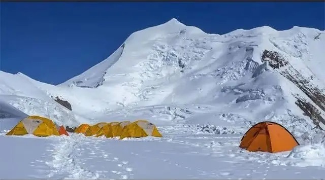 Snowy base camp with bright orange tents at the foot of towering, snow-covered mountains under a clear blue sky