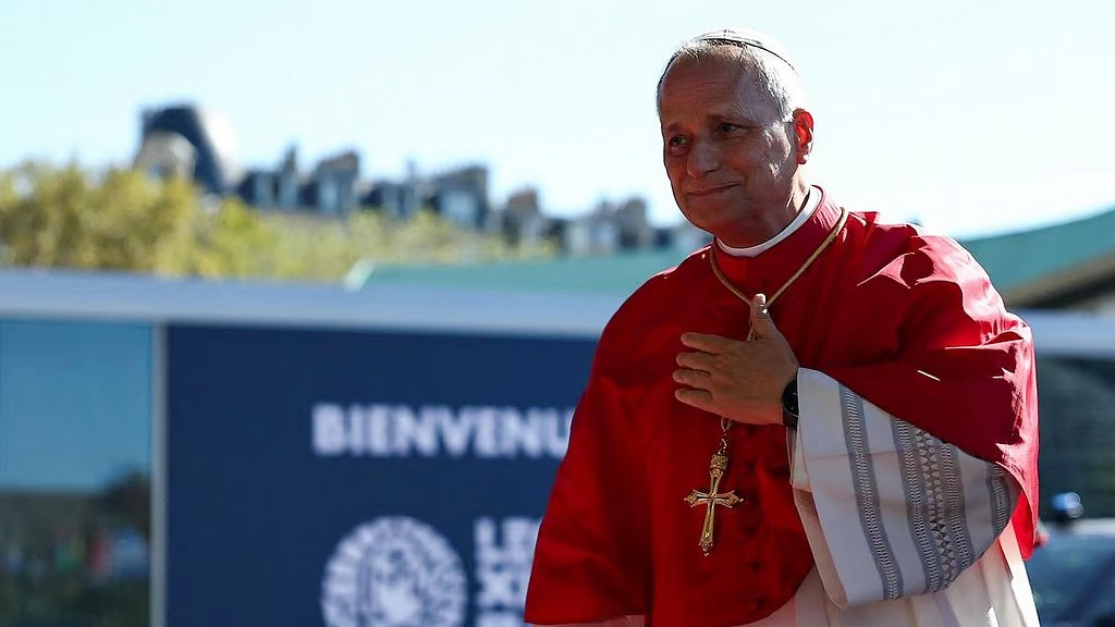 Cardinal in red vestments with a cross necklace placing a hand over his chest outdoors, near a welcome sign.