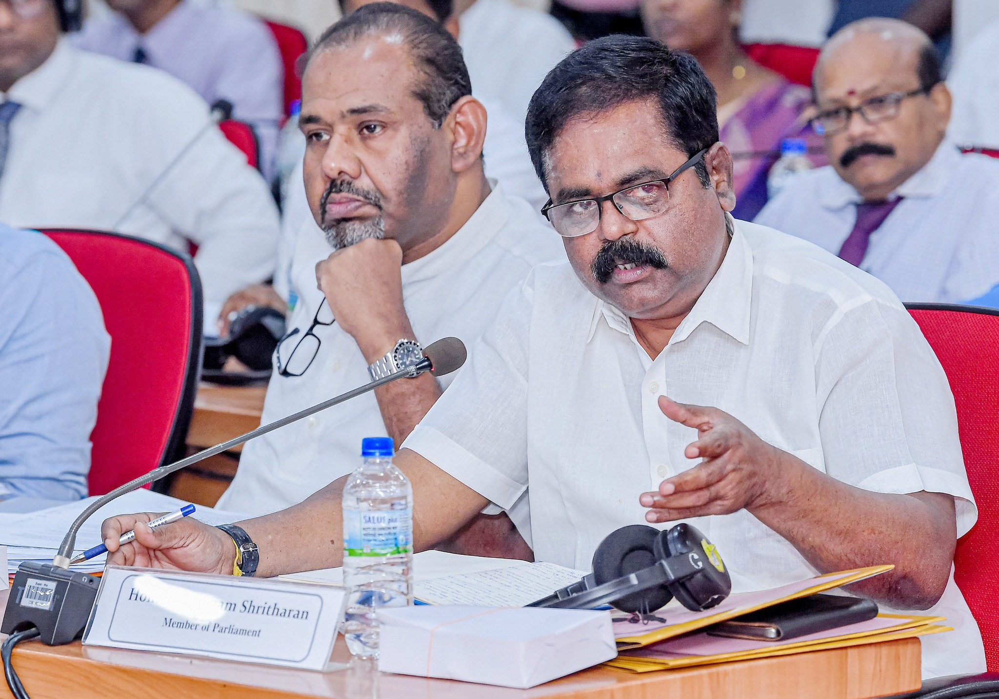 Man in a white shirt speaks into a microphone at a panel, with papers and a water bottle on the table in front of him.