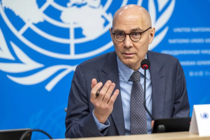 A bald man with glasses in a suit speaks at a microphone during a press conference at the UN backdrop.
