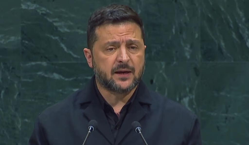 Man with dark hair and beard speaking at a podium with two microphones in front of a green marble wall.