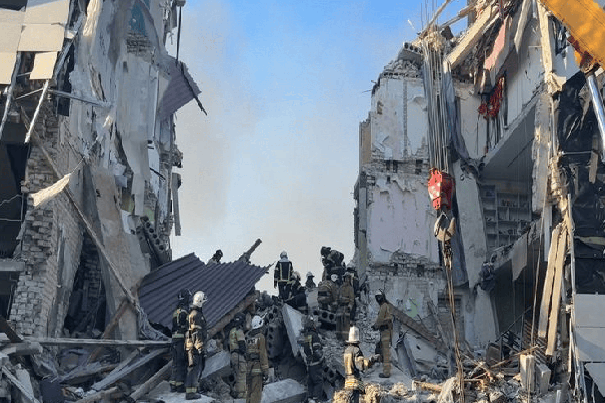 Firefighters in protective gear search through rubble between collapsed buildings after a disaster, with smoke in the background.